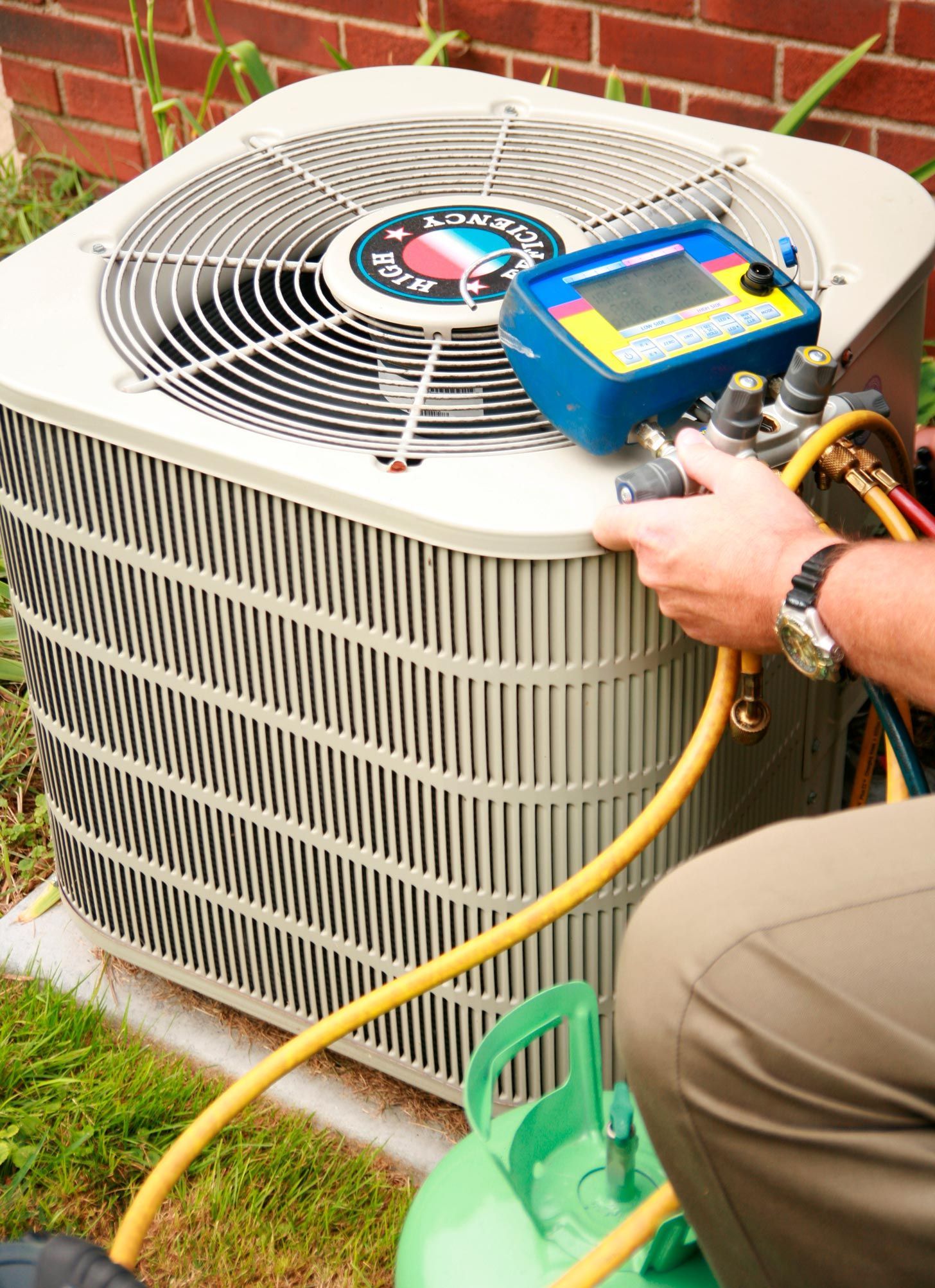 Air conditioner being serviced outdoors; a person connects gauges and a refrigerant tank.