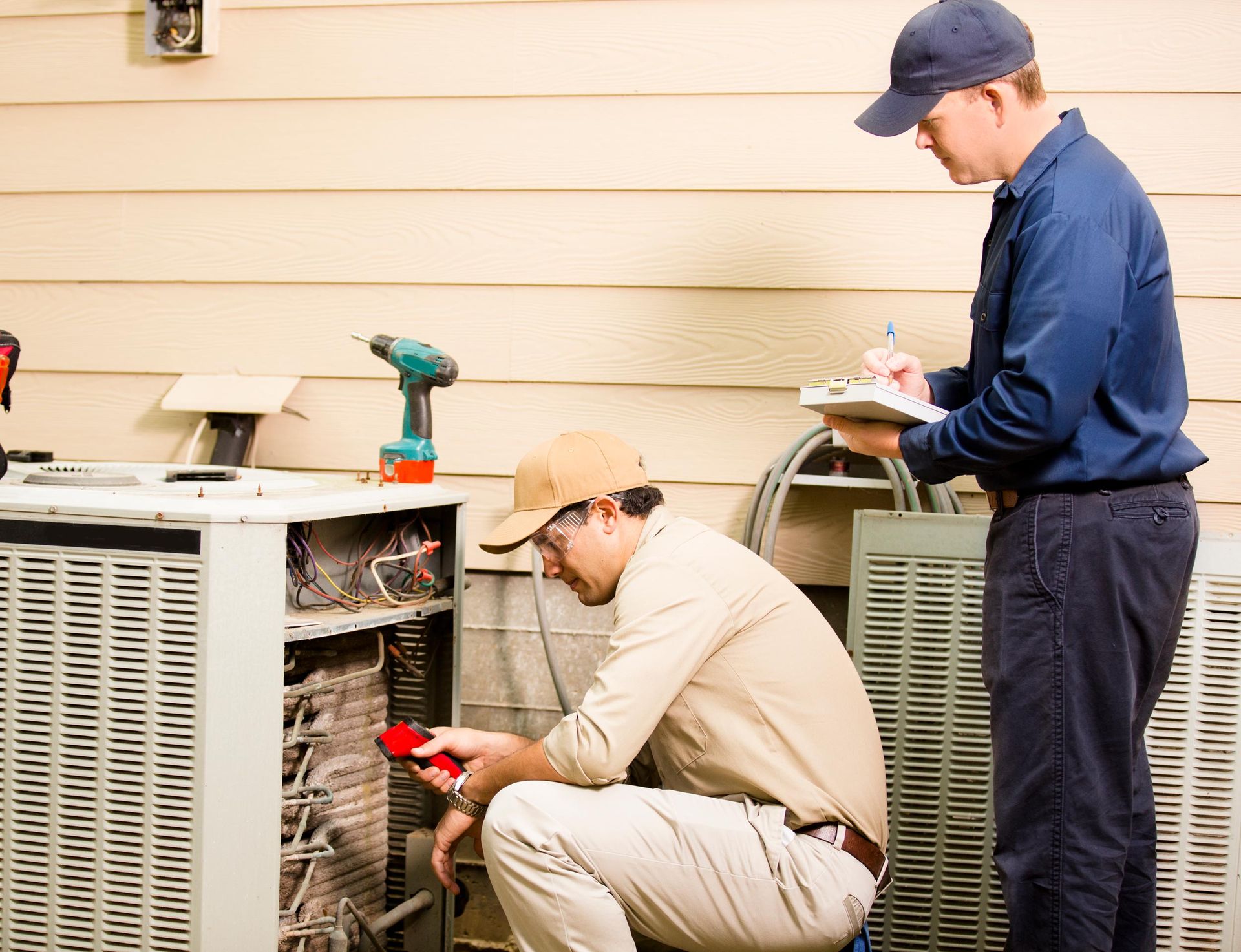 Two HVAC technicians inspecting an outdoor air conditioning unit. One checks wiring with a meter.