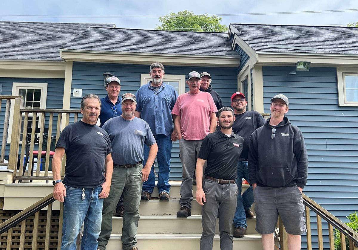 Group of ten men standing on steps in front of a blue building, some wearing hats, smiles.