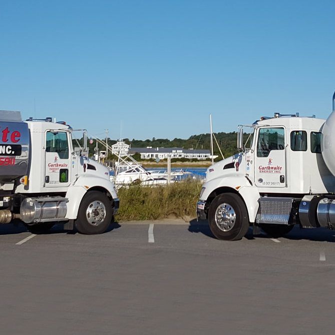 Two white tanker trucks parked side-by-side; a harbor and boats in the background on a sunny day.