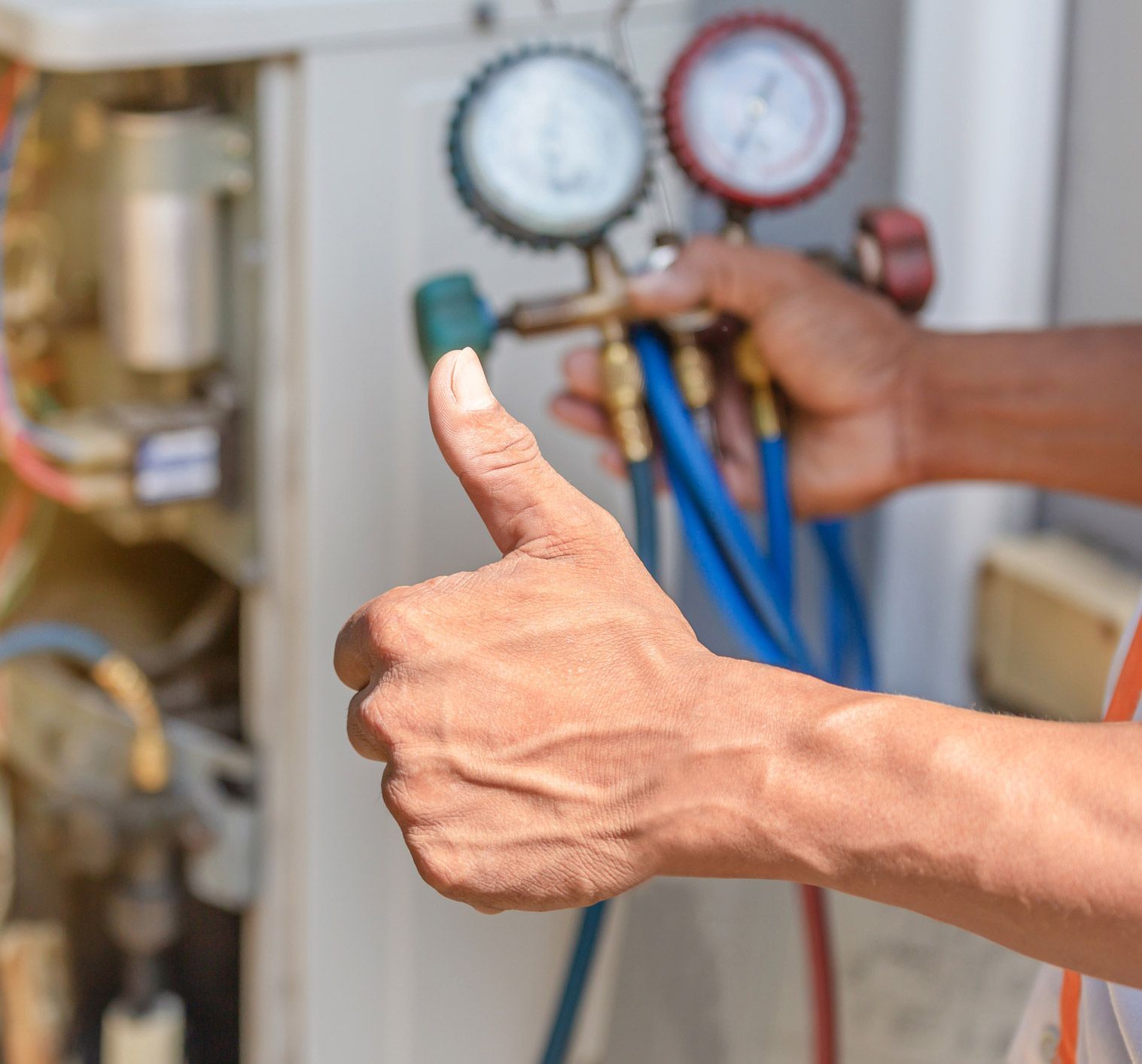 HVAC technician holding gauges, giving a thumbs up. Blue hoses, mechanical components visible.