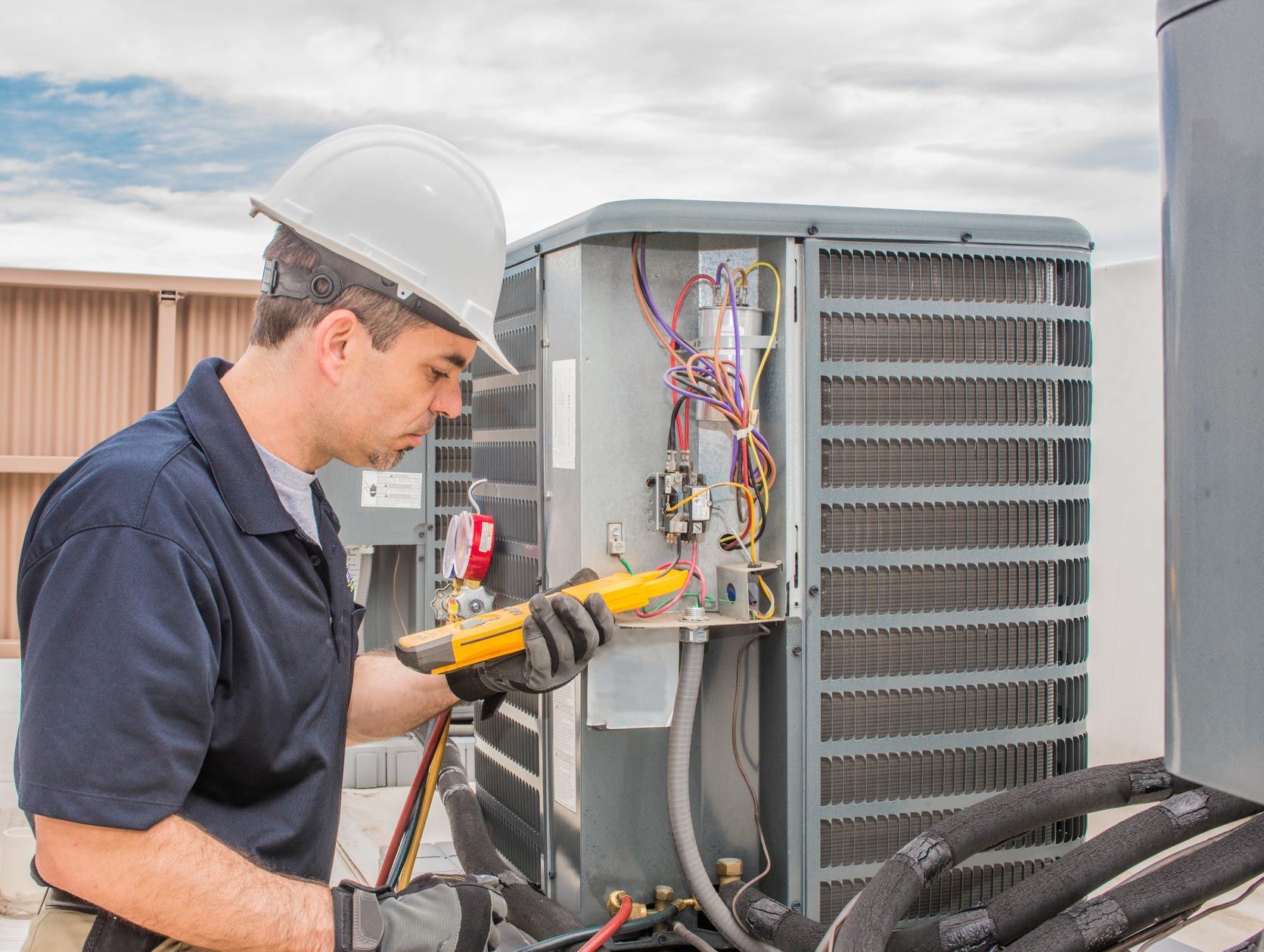 HVAC technician in white hard hat examines AC unit with a multimeter on a rooftop.