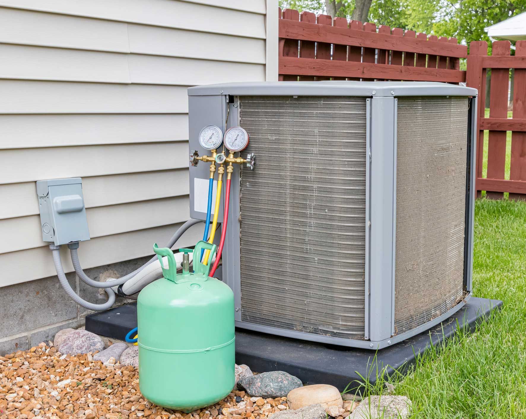 An air conditioning unit being serviced with gauges and a refrigerant tank outside a house.