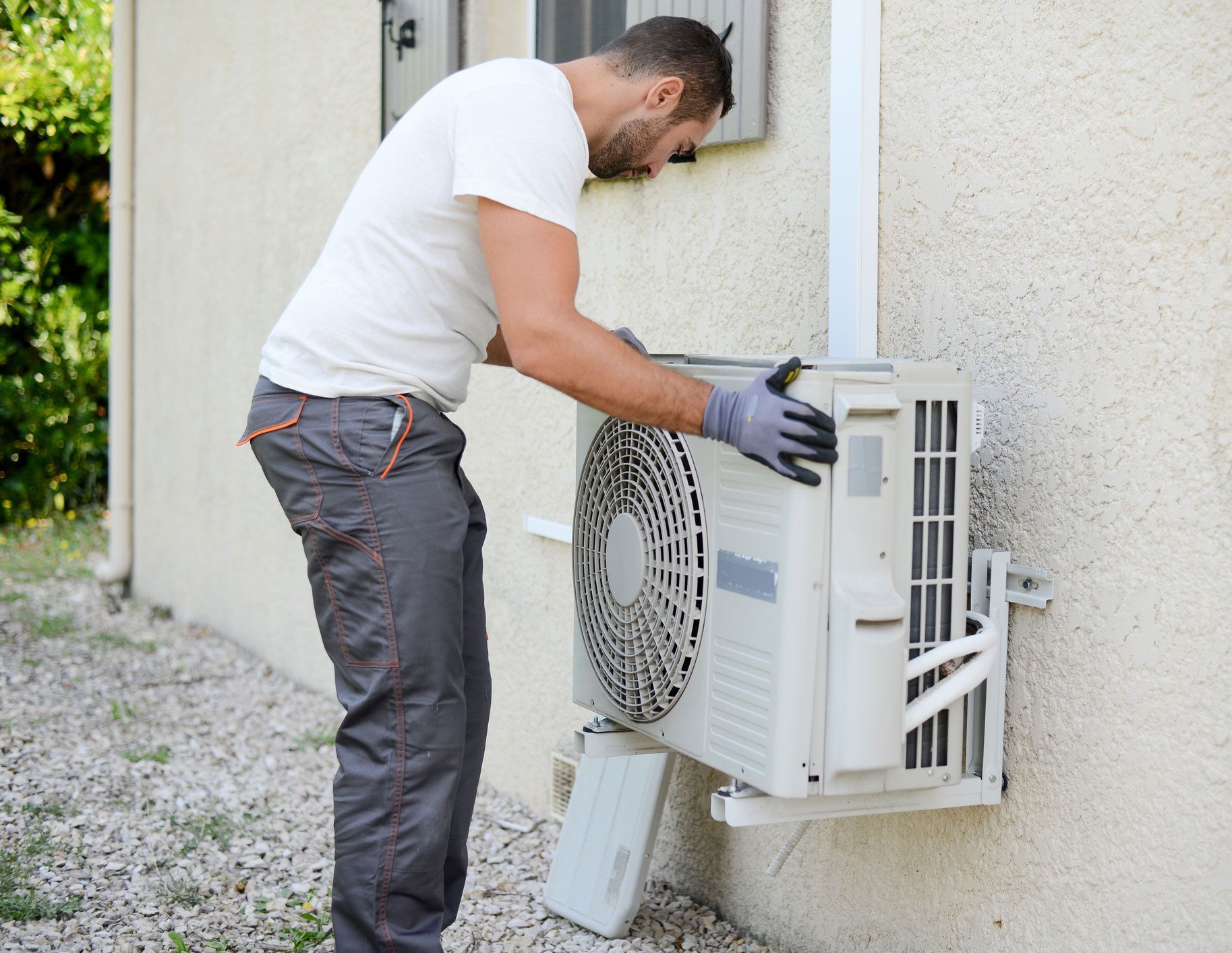 Man in gray work pants and gloves installs an air conditioning unit on a building.