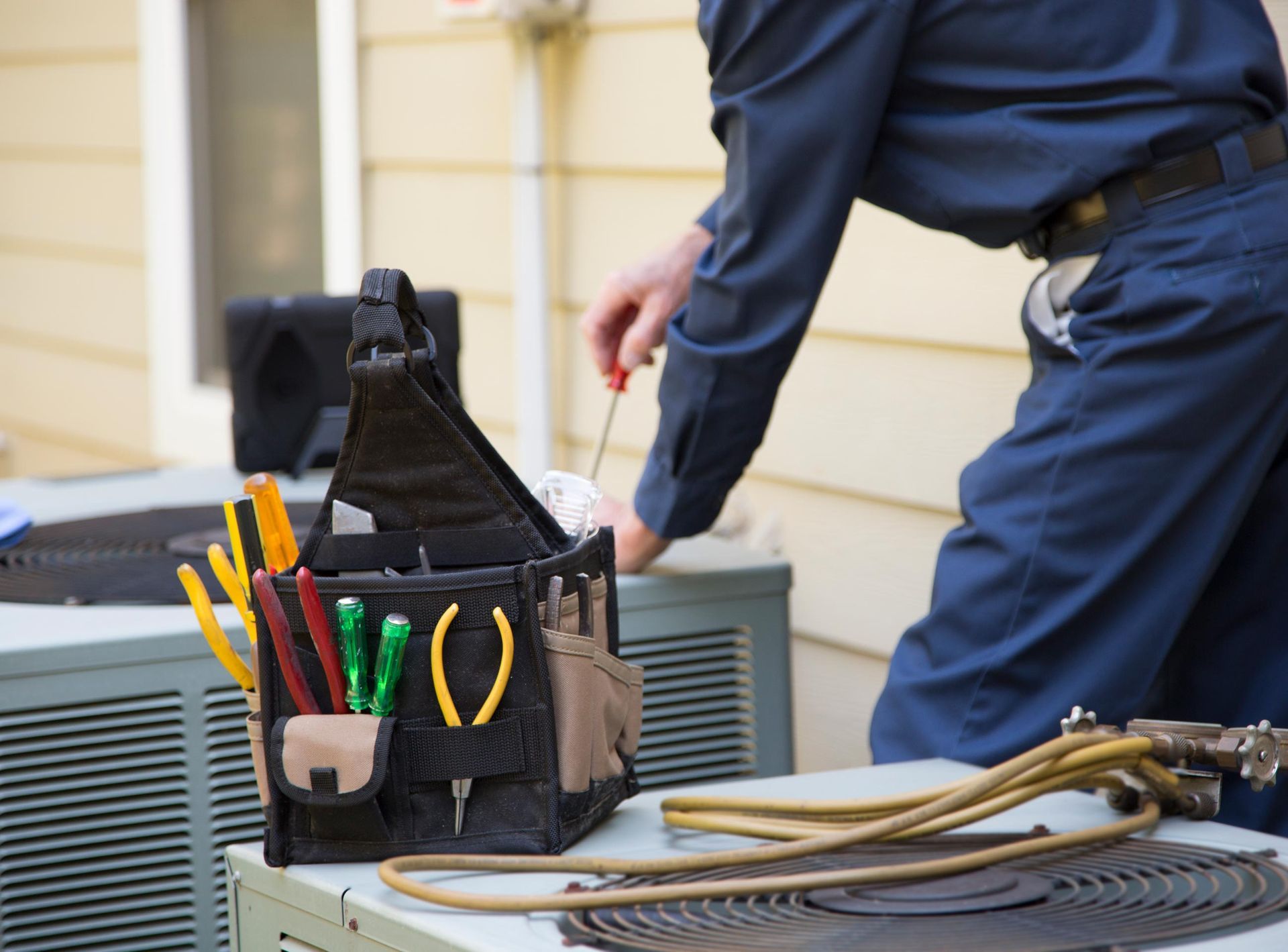 HVAC technician in blue uniform using a screwdriver on an air conditioner with a toolbox.