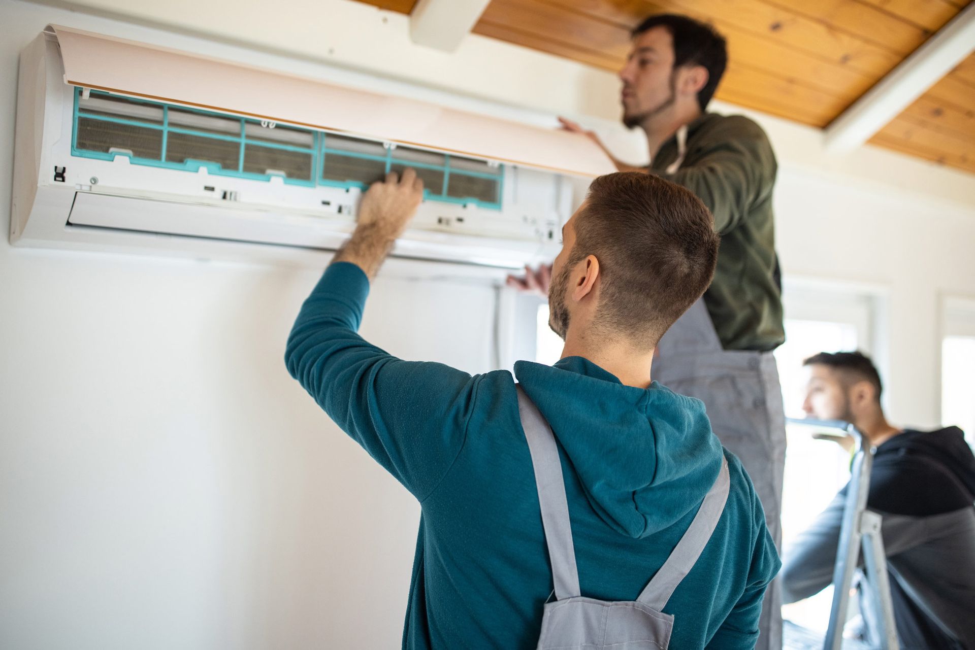 Three workers cleaning an air conditioner unit mounted on a white wall; one on a ladder, two at the unit.