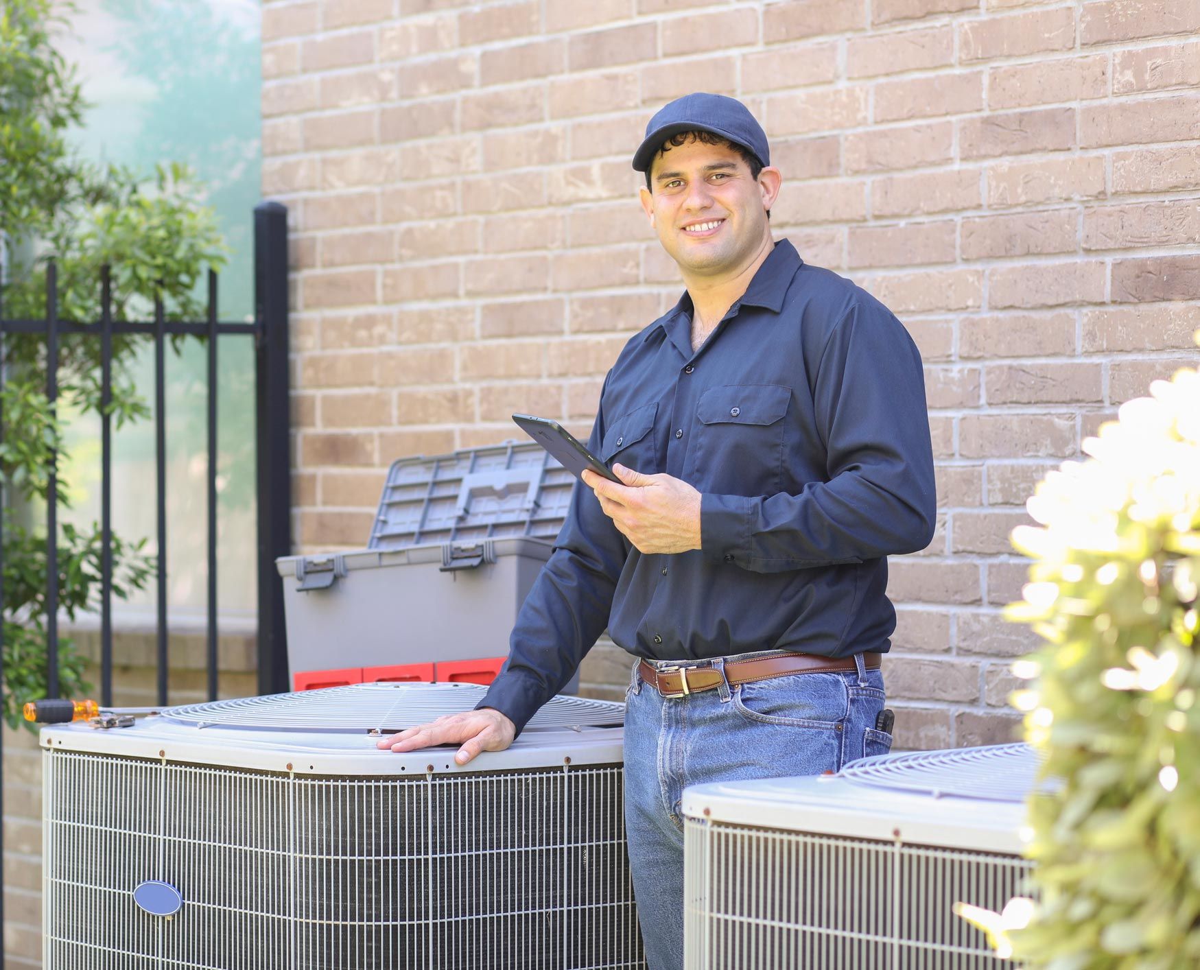 HVAC technician near air conditioning units, holding tablet, smiling in front of a brick wall.