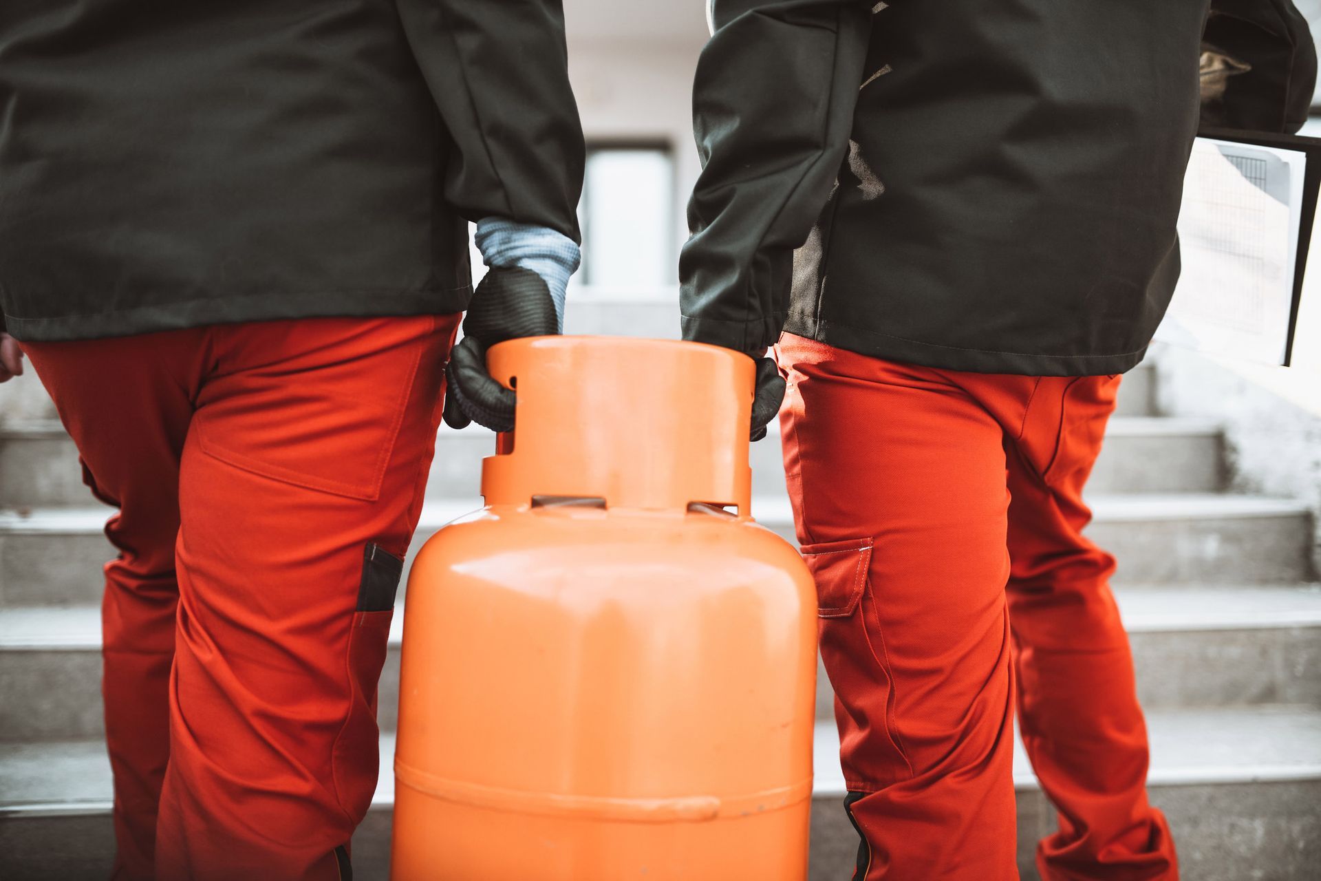 Two people in orange pants and black jackets carry an orange propane tank up stairs.