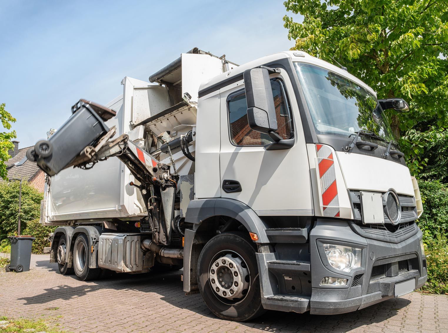 White garbage truck with a lifted arm emptying a trash bin on a driveway.