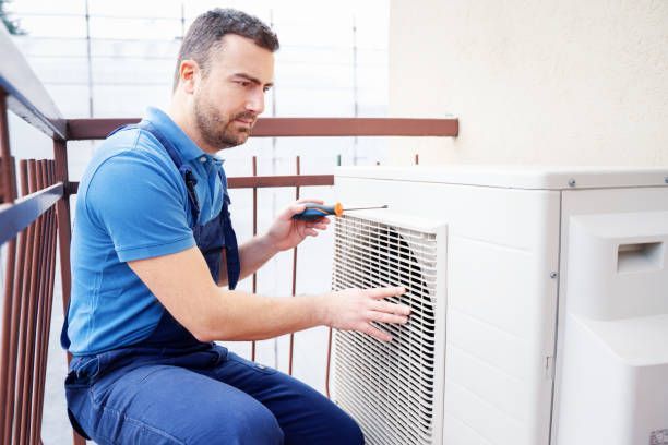 Technician in blue work clothes using a screwdriver on an air conditioning unit outside.
