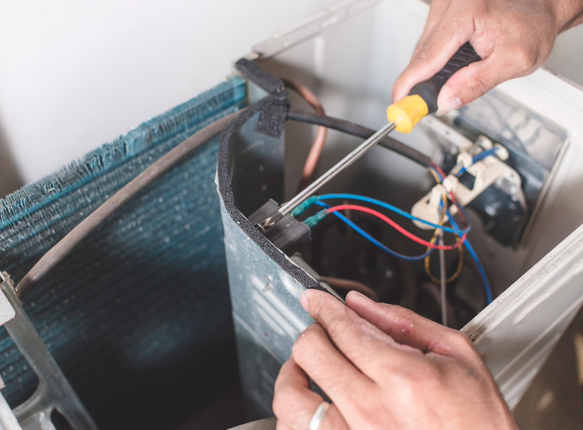 Person using a screwdriver to repair an air conditioning unit; hands close up with blue, red, and white wires visible.