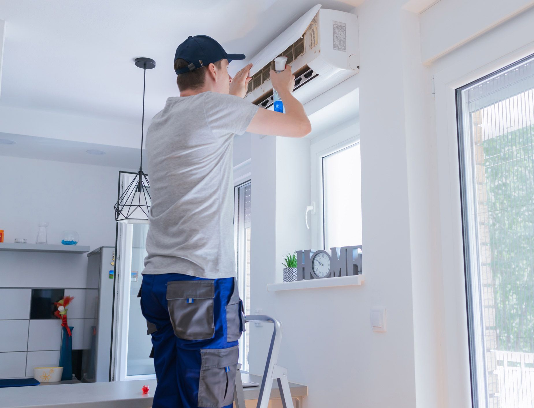 Man installing an air conditioning unit on a white wall near a window. He is wearing work clothes.