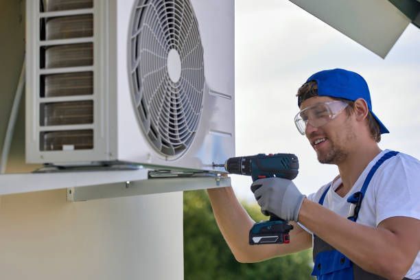 HVAC technician in blue overalls uses a drill to install an air conditioning unit. Outdoors.