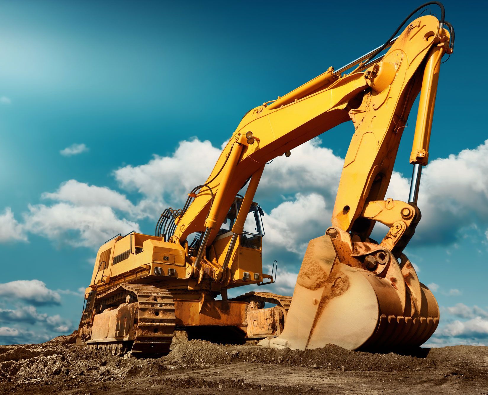 Yellow excavator on dirt against a blue cloudy sky.