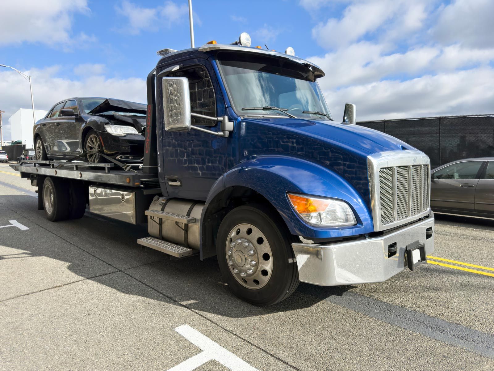 Blue tow truck with a damaged black car on its flatbed, outdoors under a blue sky.