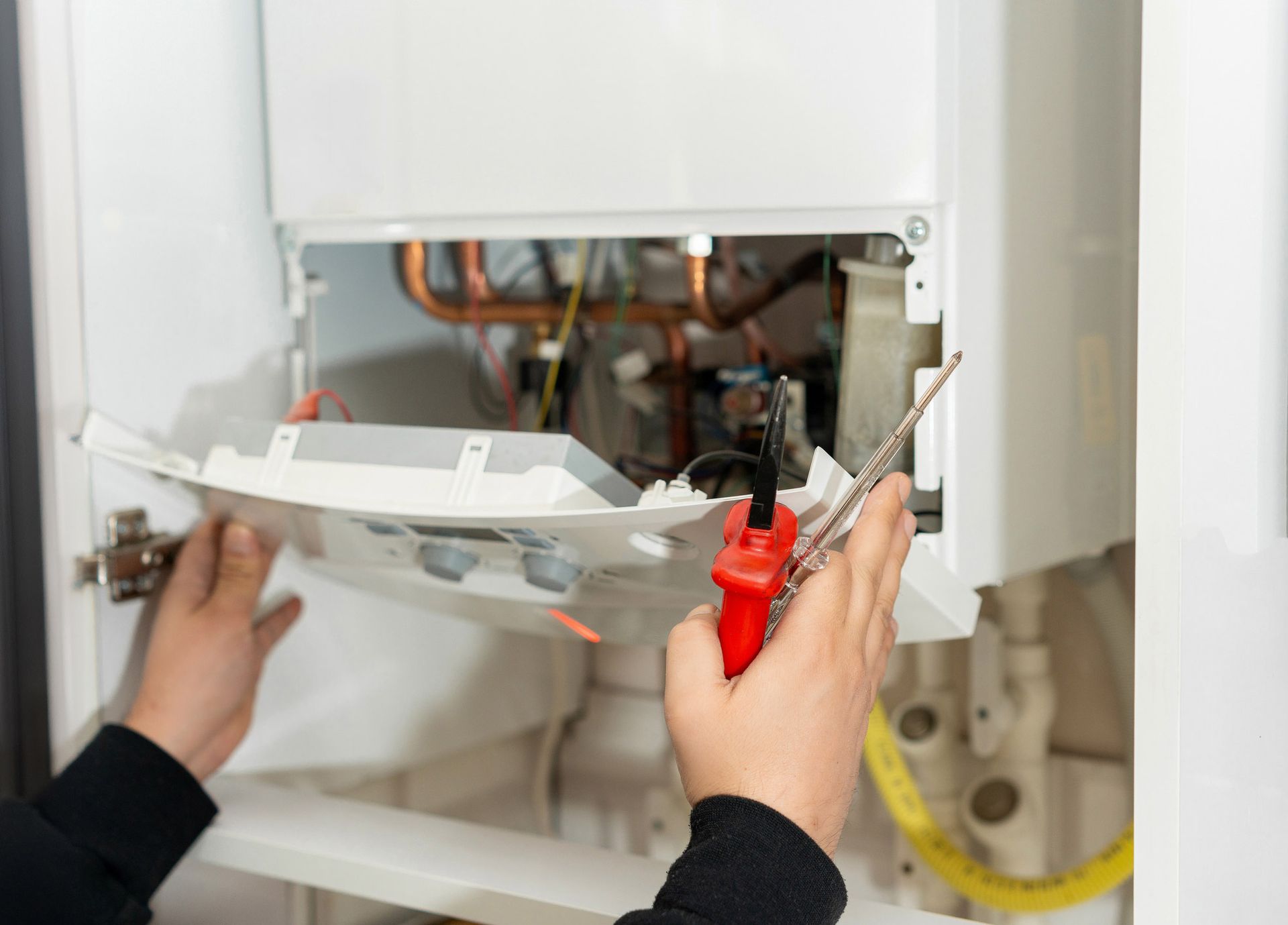 Person repairing a white furnace, holding tools inside the open cabinet.