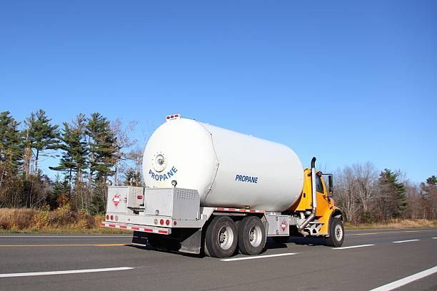 Yellow propane truck driving on a road, with blue sky and trees in the background.