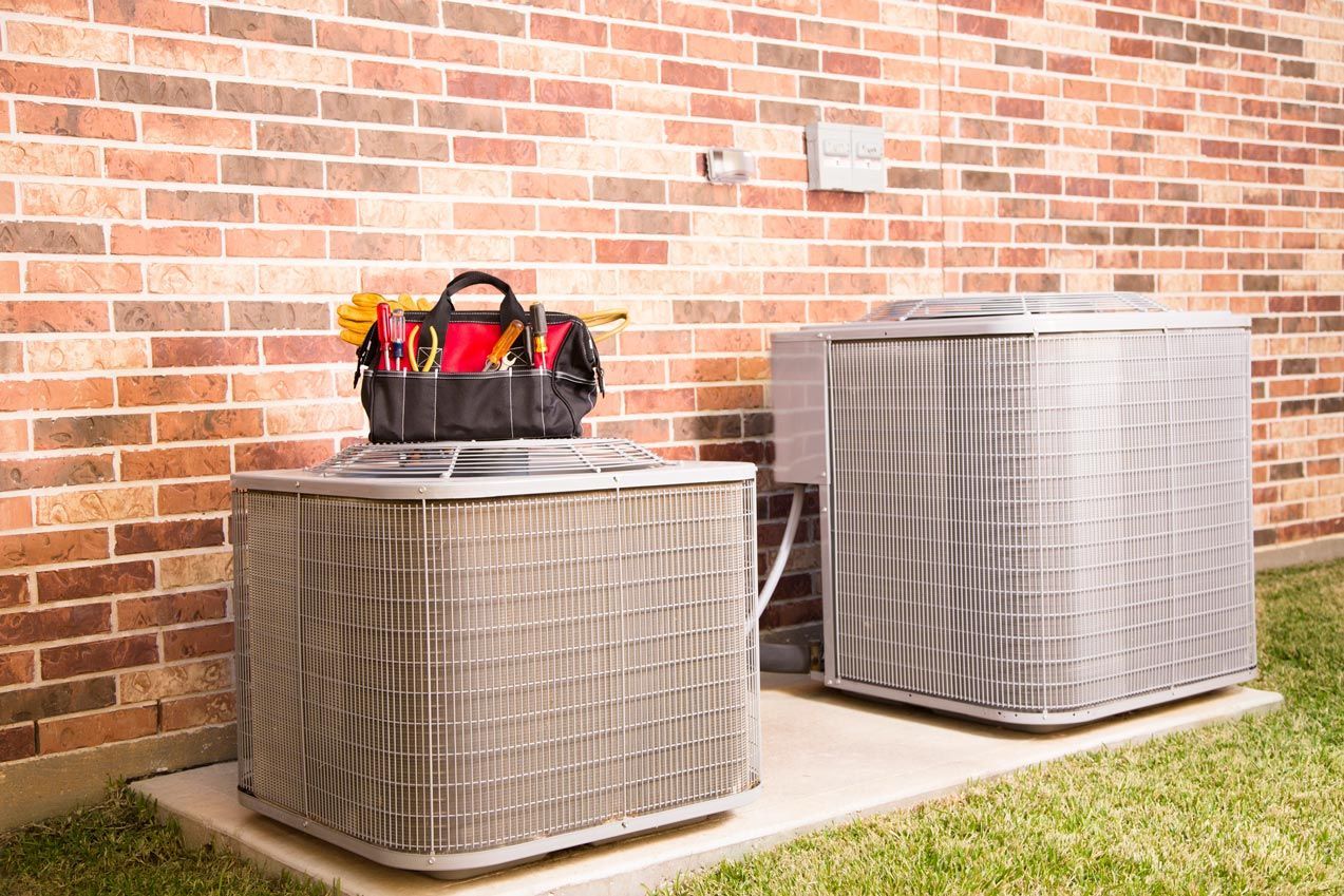 Two air conditioning units near a brick wall, with a toolbox on one.