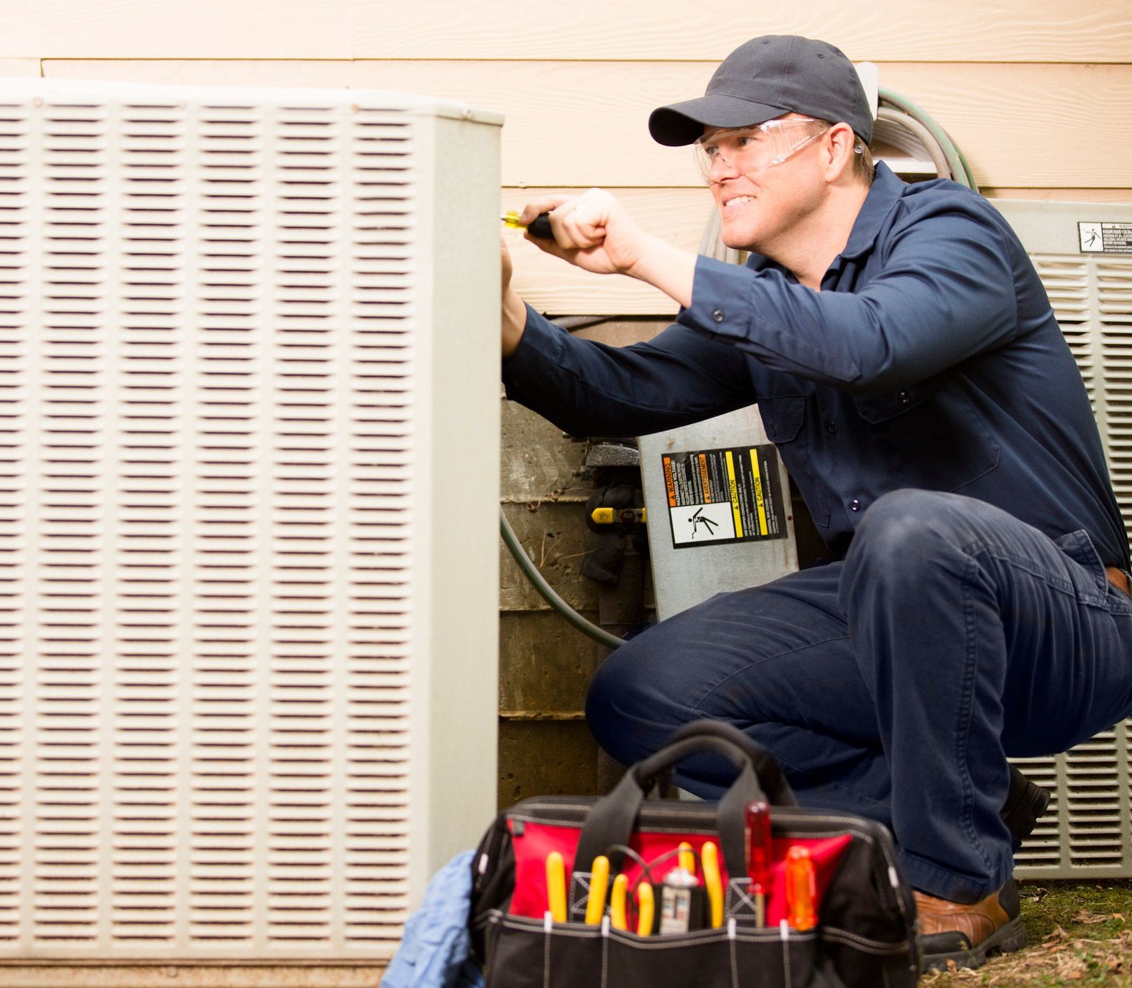 HVAC technician in blue jumpsuit, working on an air conditioner unit, tools in a bag, outdoors.
