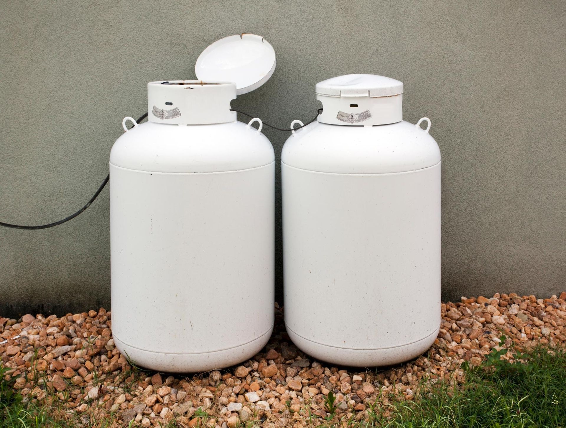 Two white propane tanks against a gray wall, on a bed of small rocks.