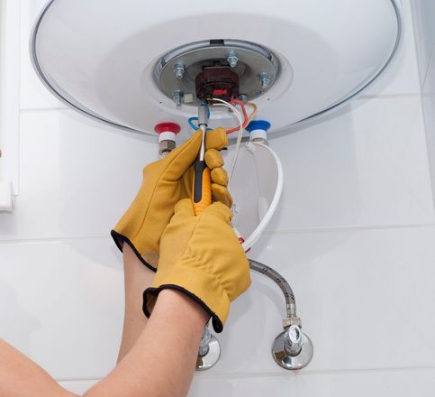 Person wearing gloves, using screwdriver on water heater electrical components.