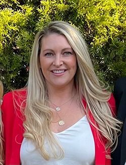 Woman with long blonde hair, smiling, wearing a red blazer over a white top, necklace.