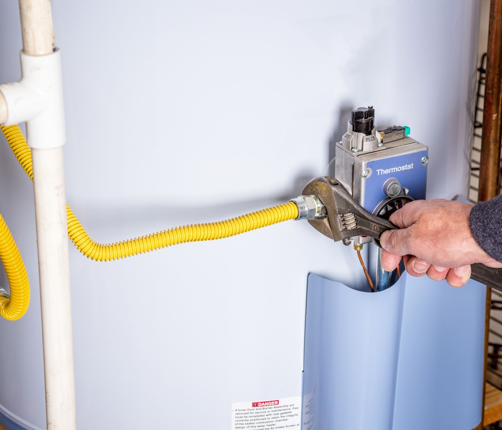 A person uses a wrench to tighten a yellow gas line connected to a water heater in a utility room.