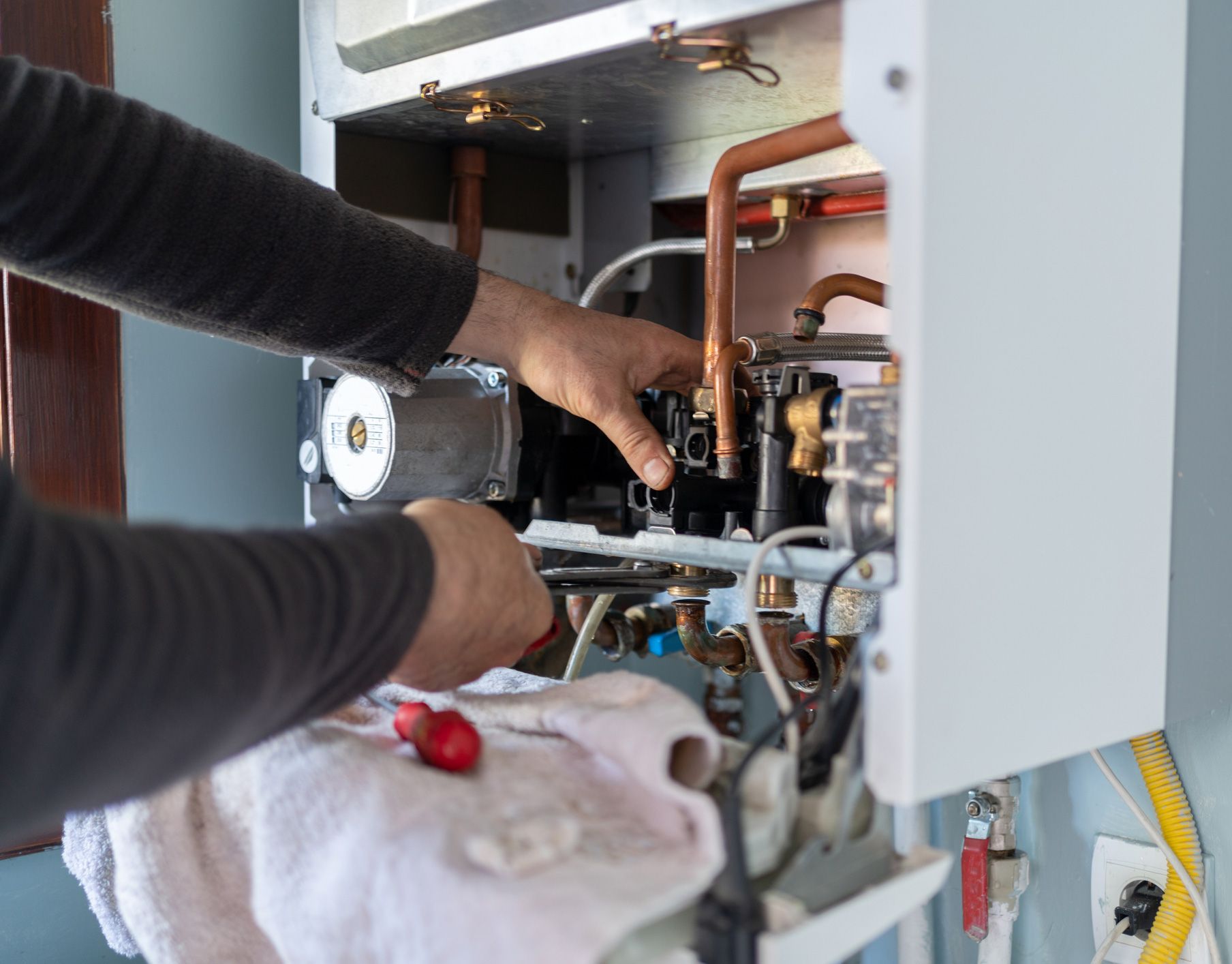 Person repairing a gas boiler; hands working on internal components; tools visible.