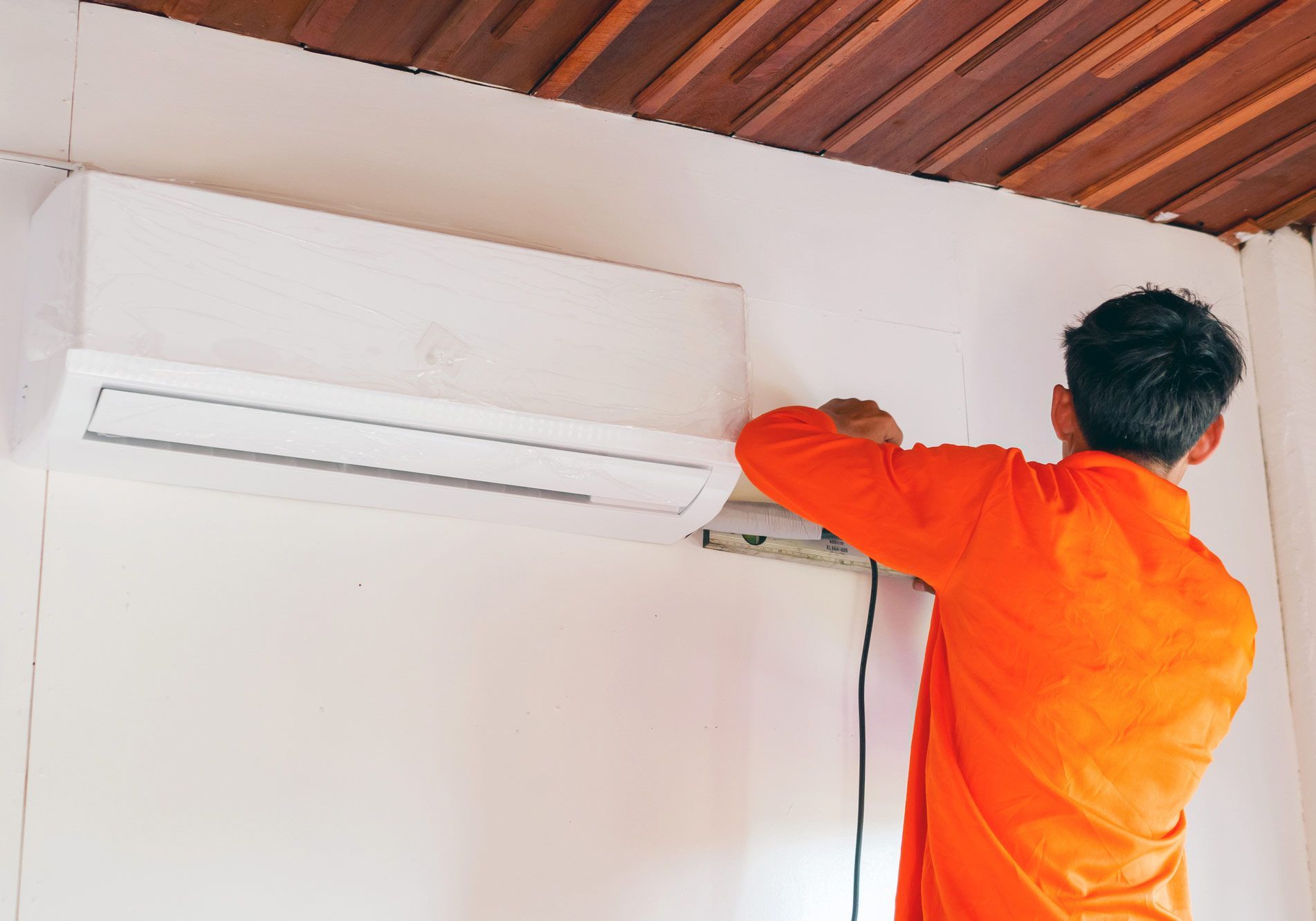 Person in orange shirt installing an air conditioner unit on a white wall, indoors.