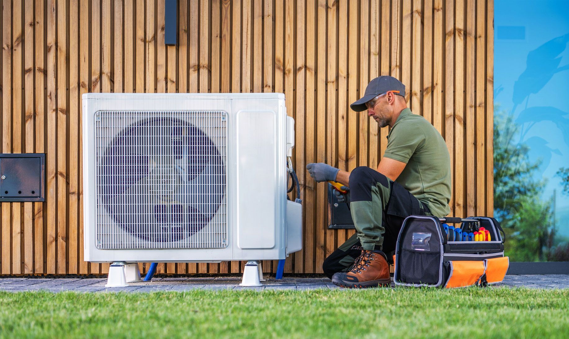 Man in work clothes working on a white heat pump, outdoor installation on a wooden building.