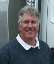Man with gray hair, wearing glasses, and a blue sweater smiles outdoors.