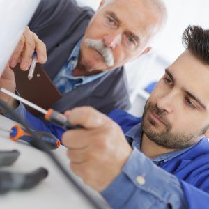 Man using a screwdriver, supervised by an older man holding a clipboard.