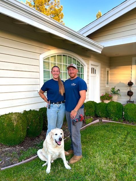 Man and woman with dog in front of a house, both wearing work shirts, standing near shrubs.