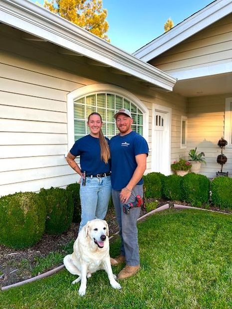 Man and woman with dog in front of a house, both wearing work shirts, standing near shrubs.