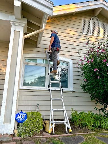 Person on a ladder, working on the side of a house. The house is light-colored with white trim and a window.