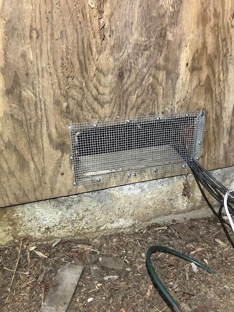 Close-up of a wooden wall with a rectangular vent covered by wire mesh. Electrical wires extend out.