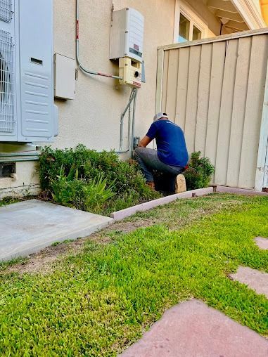 A person installing electrical equipment outdoors. They're kneeling near a wall with wires and shrubbery.