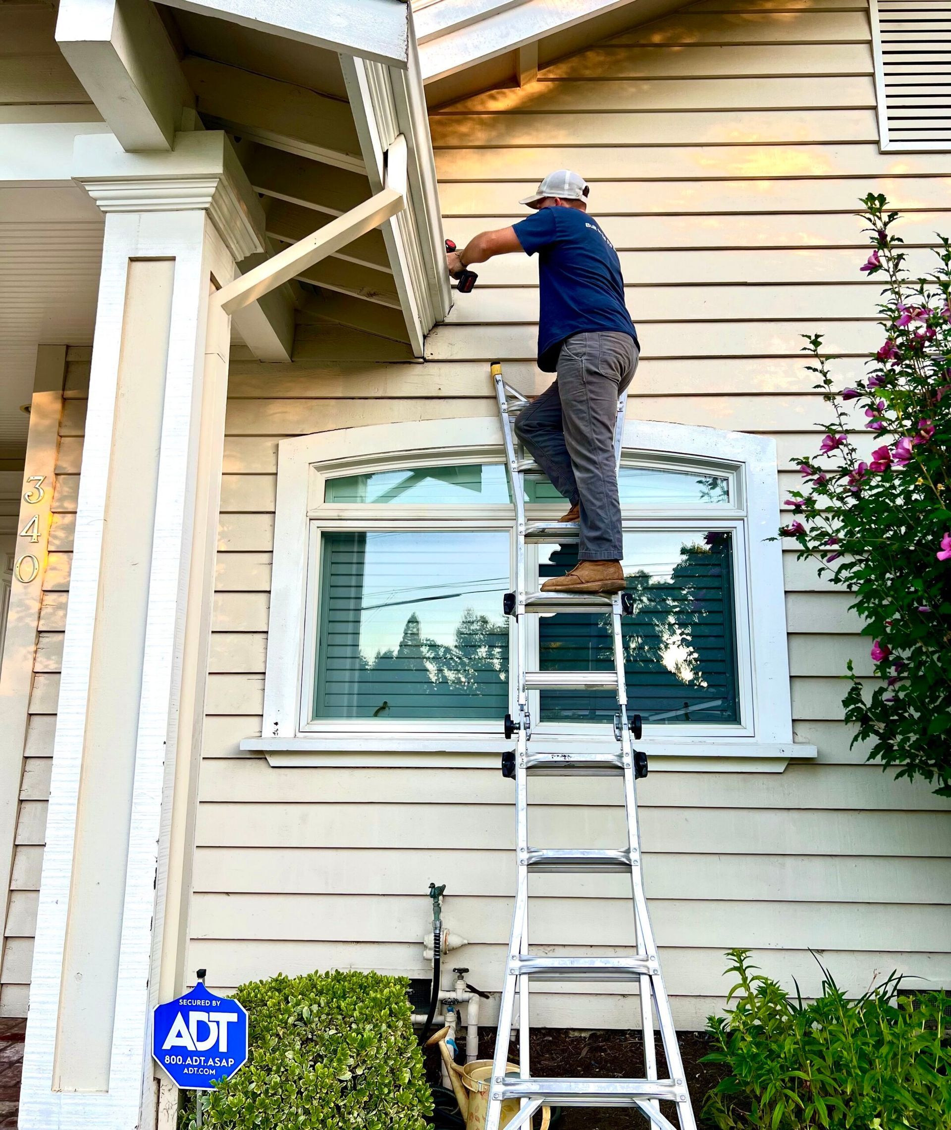 Person on ladder installing gutter on a light-colored house. Green bushes and a blue security sign are visible.