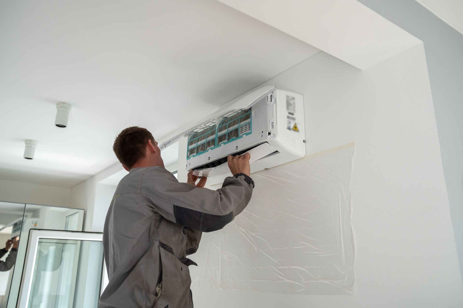 Man in gray coveralls installs an air conditioning unit on a white wall.