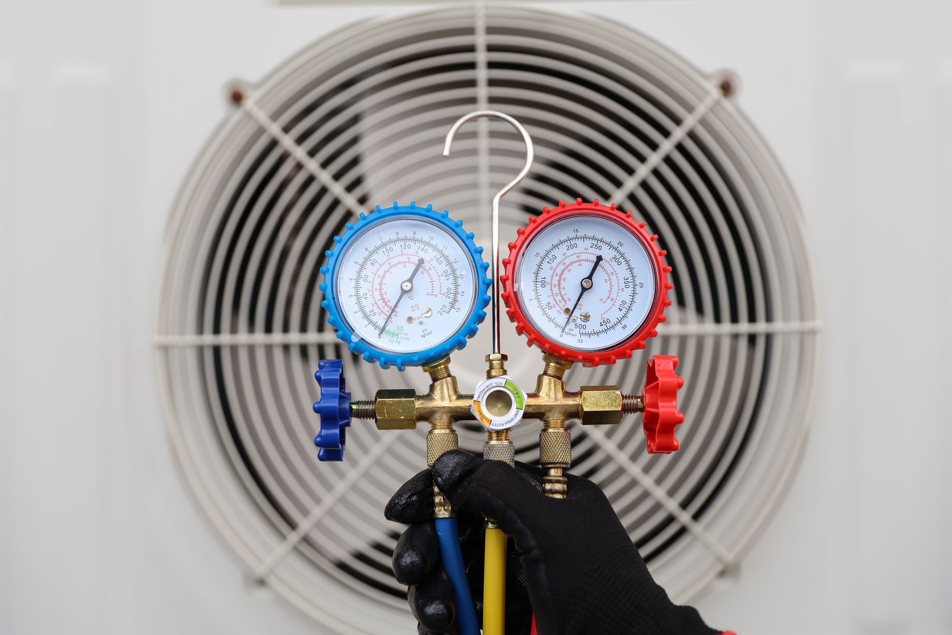 Hand holding manifold gauge set in front of an AC unit's fan, showing pressure readings with blue and red gauges.