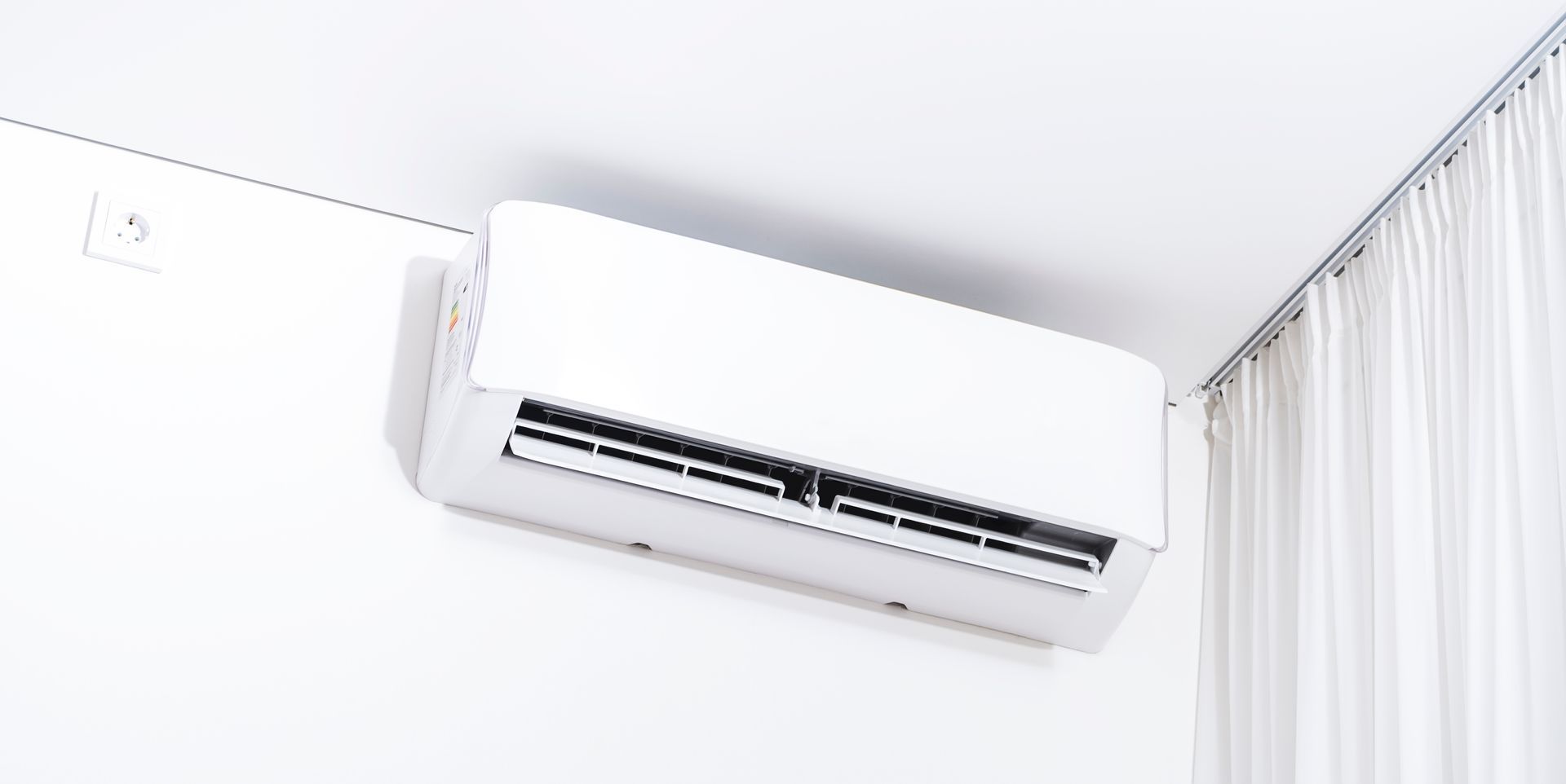 White air conditioner mounted on a white wall. White curtains in the corner.