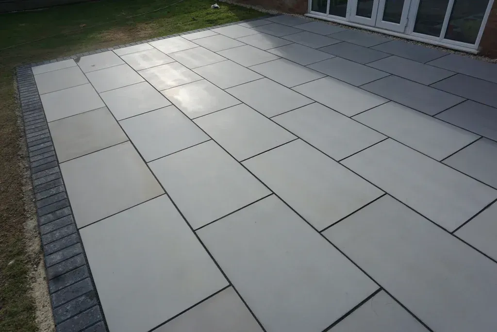 Patio with large light-gray tiles beside a lawn and house windows, viewed in sunlight
