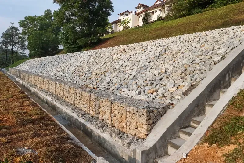Stone retaining wall on a grassy hillside beside a house, lined with concrete steps.