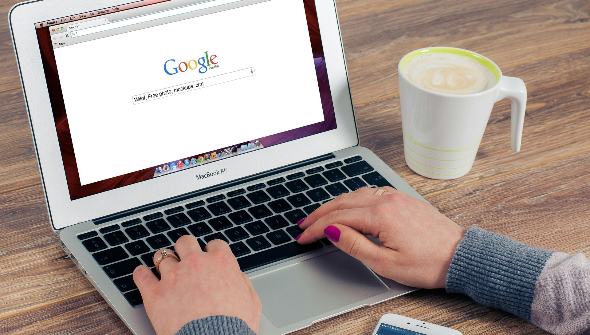 Hands typing on a laptop with Google search open, a coffee mug, and a smartphone on a wooden desk.