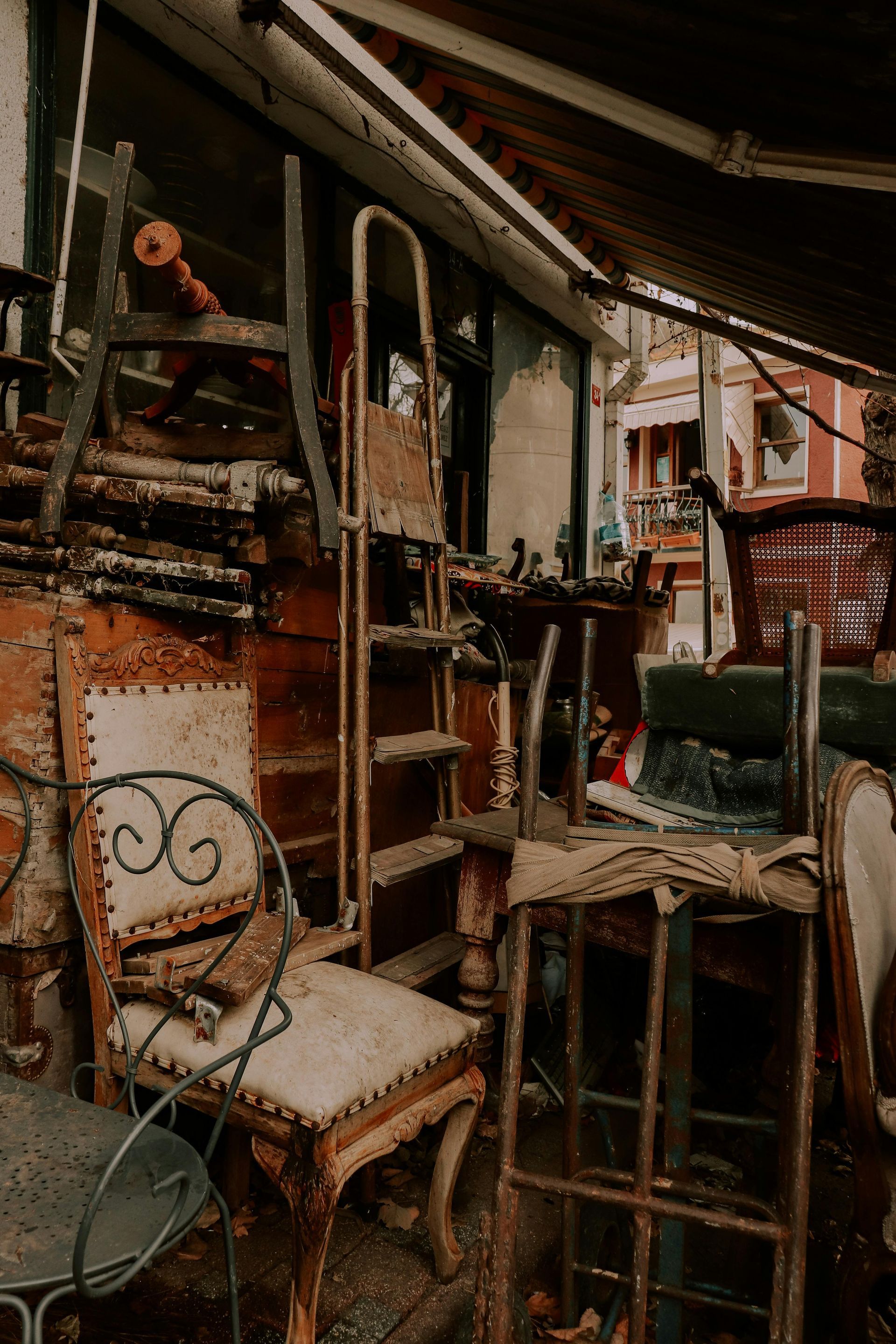 A cluttered outdoor antique shop displaying weathered chairs, ladders, and furniture against a brick building.