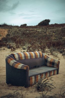 A worn sofa with orange and gray stripes sits on sand, amid dry vegetation under a cloudy sky.