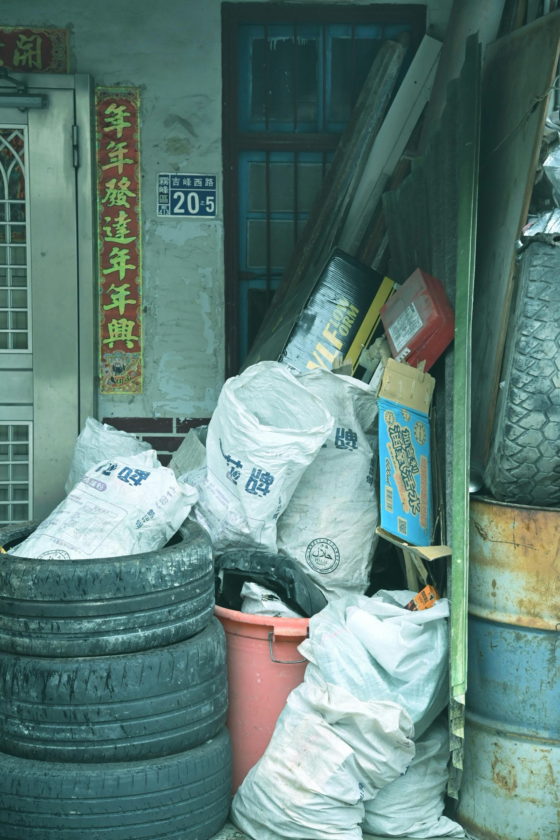 A cluttered alleyway next to a building. White bags, tires, and debris are piled against a wall with a window and a doorway.
