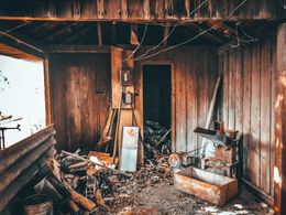 Interior of a dilapidated wooden building. Debris litters the floor; a doorway leads to darkness.