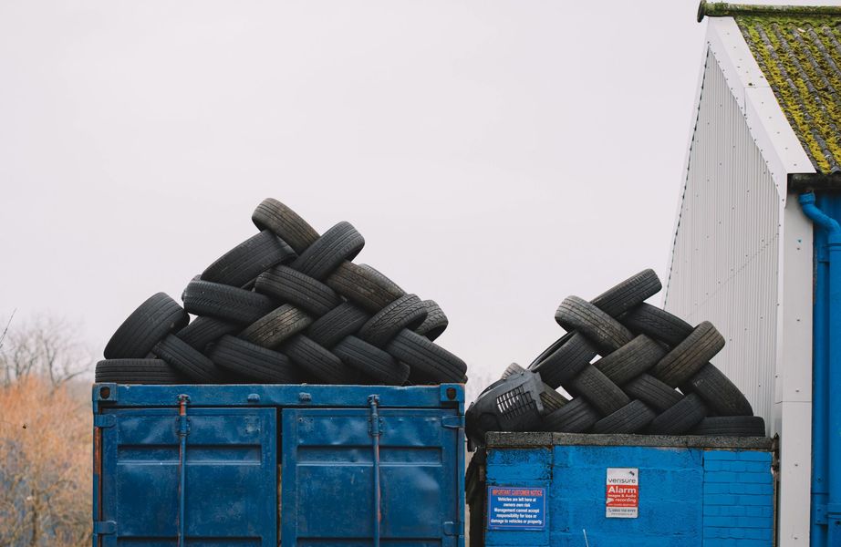 Blue containers overflowing with a pile of used tires, stacked on top. A white building is to the right.