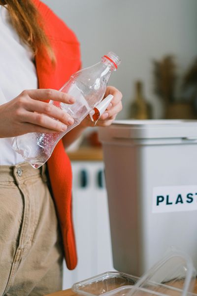 Woman putting a plastic bottle into a recycling bin. She wears a red jacket and tan pants.